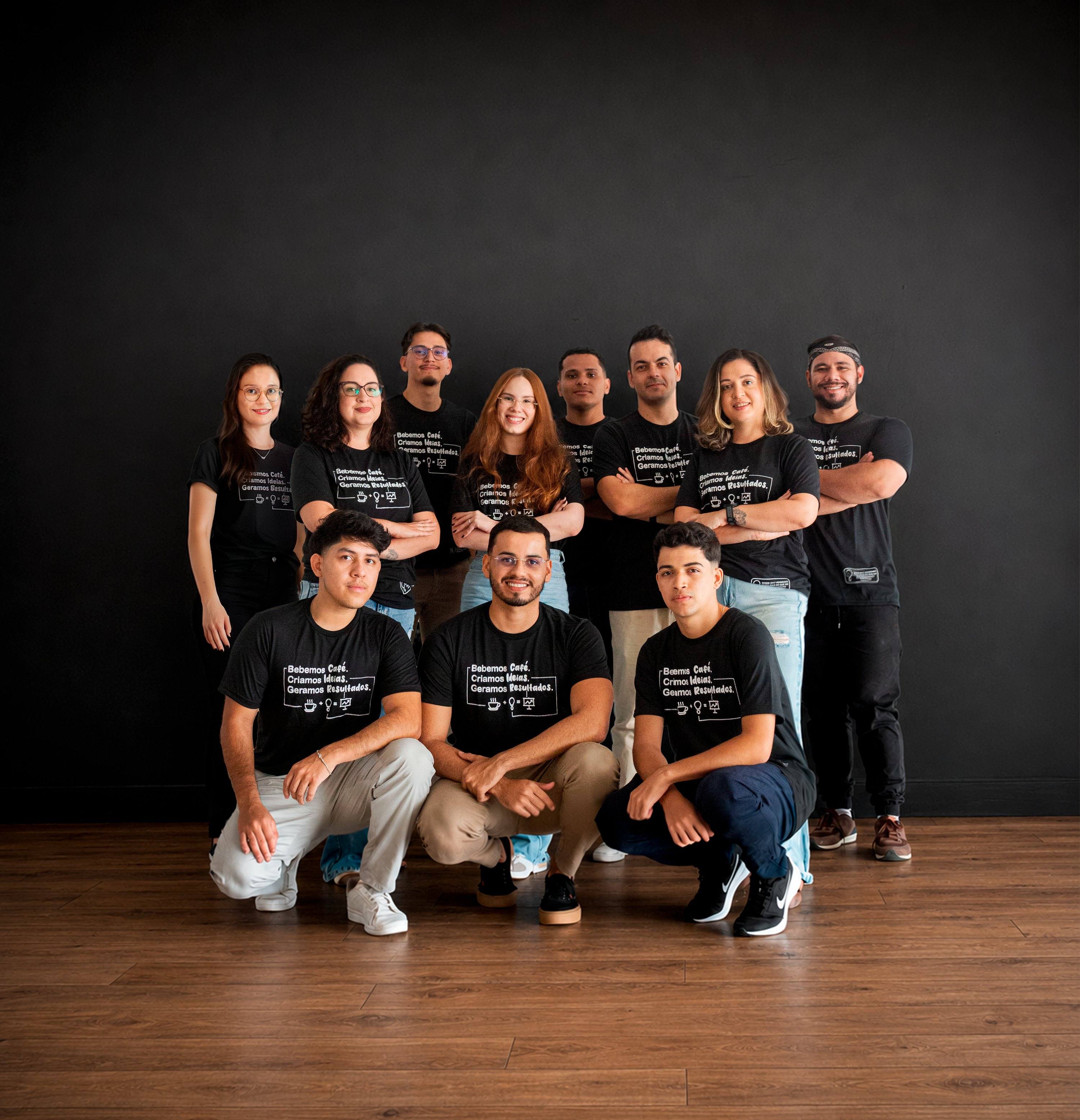 Equipe da Agência Ideia reunida em frente a uma parede preta, vestindo a camiseta com a frase “Bebemos café. Criamos ideias. Geramos resultados.”